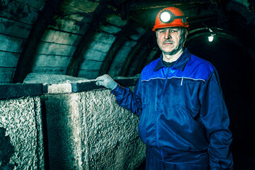 A tired miner in an old coal mine is standing near a broken trolley. A miner in a blue protective...
