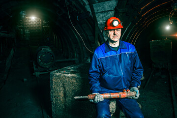 A tired miner in an old coal mine sits on a broken trolley. A miner in a blue protective suit with...