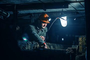 Tired young miner in an old coal mine. A miner in a protective suit with a helmet on his head and a flashlight works in the dark. Work in a coal mine.