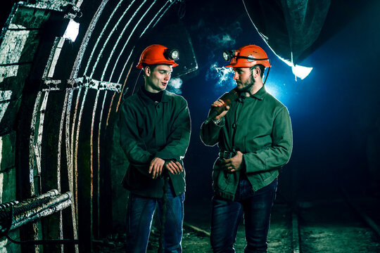 Two tired young miners in an old coal mine communicate with each other at work. Miners in a protective suit with a helmet on their heads and a flashlight stand in a cold tunnel.