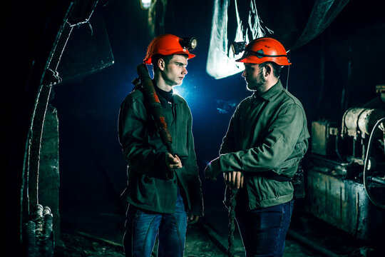 Two tired young miners in an old coal mine communicate with each other at work. Miners in a protective suit with a helmet on their heads and a flashlight stand in a cold tunnel.