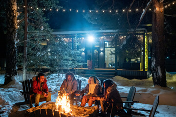 Group of friends sitting near the fire outdoors, cooking marshmallows and talking till late evening during winter vacations in the country house