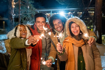 Portrait of happy friends smiling at camera standing outdoors with sparklers in their hands and...
