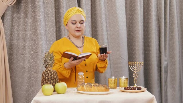 A Jewish woman in a traditional headdress reads kiddush for wine during a Shabbat meeting. Overall plan
