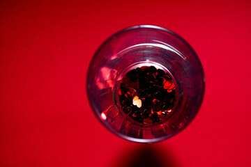 Wedding ring and confetti of their red hearts in a champagne glass on a red background. Top of view