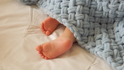 The legs of a sleeping child peeks out from under a blue plaid. Blue plaid knitted by hand. Home...