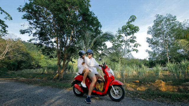 Love couple on red motorbike in white clothes on forest road trail trip. Two caucasian tourist woman and man kiss hugs sit on scooter. Motorcycle rent Safety helmet sunglasses. Asia Thailand tourism.
