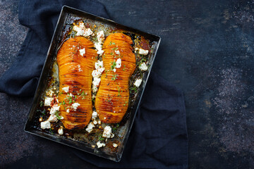 Traditional fried hasselback butternut squash pumpkin roast with herbs and feta sheep cheese served as top view in a rustic metal tray on an old black board with copy space right - obrazy, fototapety, plakaty