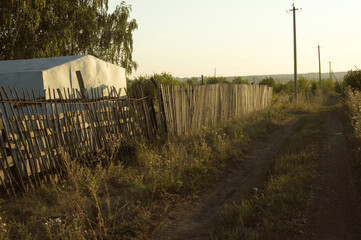 Summer rural evening landscape with a road, wooden fence, metal barn, birch, electric poles at sunset
