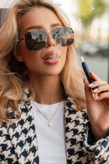 Stylish beautiful young blonde woman with sunglasses and sexy lips smokes an e-cigarette and blows...