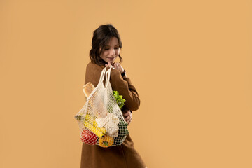 Back view of happy smiling young woman in brown dress holding reusable string bag with organic fruits and vegetables over orange background. Sustainability, eco living and people concept.