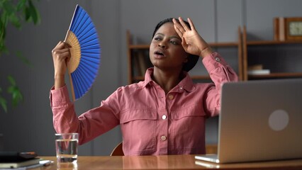Sweaty young african american woman employee sit by laptop at hot office try to avoid overheating use paper fan. Sad stressed female manager freelancer tired of work in uncomfortable room breath hard 