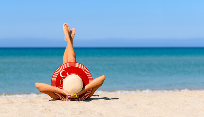 A slender tanned girl on the beach in a straw hat in the colors of the flag of Turkey. The concept...