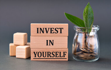 INVEST IN YOURSELF - words on wooden blocks with a jar of coins on a gray background