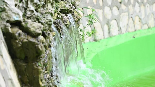 Fountain and flowing water closeup. Flowing water between river stones in natural lagoon. Small pond with fountain. Source of crystal water and drops reflect sun's rays. Pure aqua in movement. Park an