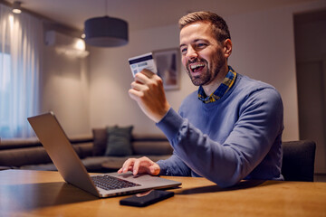 An excited man getting new credit card and spending money online on the laptop from home.