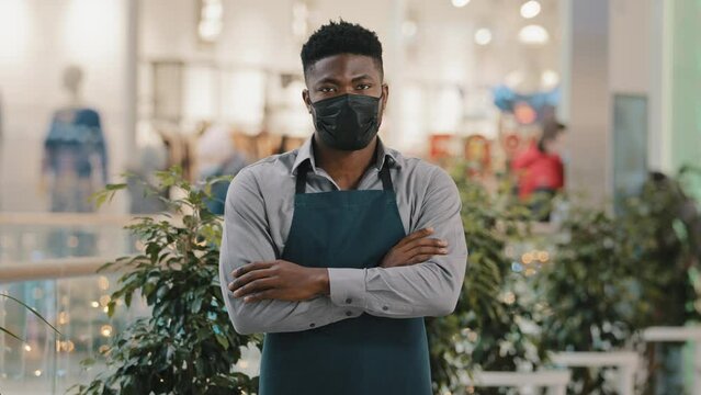 Confident african american man wearing medical mask standing indoors looking at camera young waiter in barista apron employee cafe bar owner seller posing with arms crossed small business concept