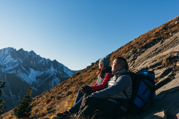 Fototapeta premium Man and woman in sportswear sit on the hill. Lovely couple of travelers to hold, enjoying highland landscape, beautiful sunset. Two tourists are hiking against the backdrop of snow-capped mountains.