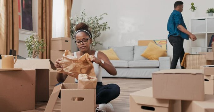 Beautiful woman with glasses sits on floor of purchased rented apartment unpacks cardboard boxes of stuff takes out vase wrapped in paper smiles at camera in background man is cleaning