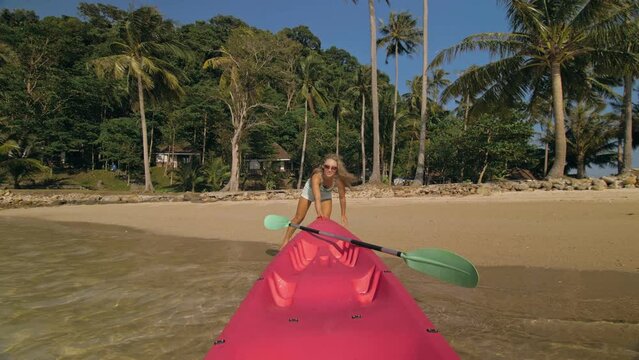 Young woman and sunglasses pulls pink plastic canoe with paddle to azure sea water from beach with palms. The beginning of the boat trip, boarding. Traveling to tropical countries