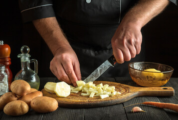 The chef cuts raw potatoes into small pieces with a knife for French fries. Close-up of a cook hands while working in a kitchen