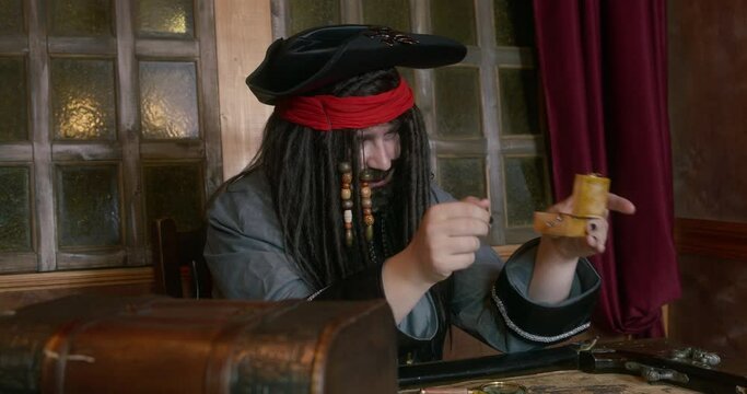 Pirate captain in cocked hat with long hair twists stolen musical box sitting AT table near treasure chest against window in ship cabin