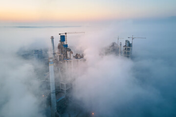 Fototapeta premium Aerial view of cement factory with high concrete plant structure and tower crane at industrial manufacturing site on foggy evening. Production and global industry concept