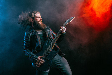 Expressive man rock guitar player with long hair and beard plays on the smoke background. Studio...