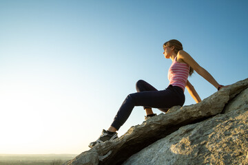 Woman hiker sitting on a steep big rock enjoying warm summer day. Young female climber resting during sports activity in nature. Active recreation in nature concept.