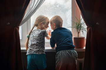Children, a boy and a girl look out the window of their house during a snow storm