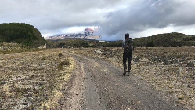 A tall man is walking on the moorlands of Sincholagua in the Andes of Ecuador. He is walking on a trail path toward the mountain. Clouds are seen above the hills.