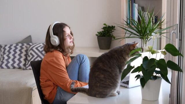 Relaxed teenage girl listening to music on headphones and looking out of the window with her cat