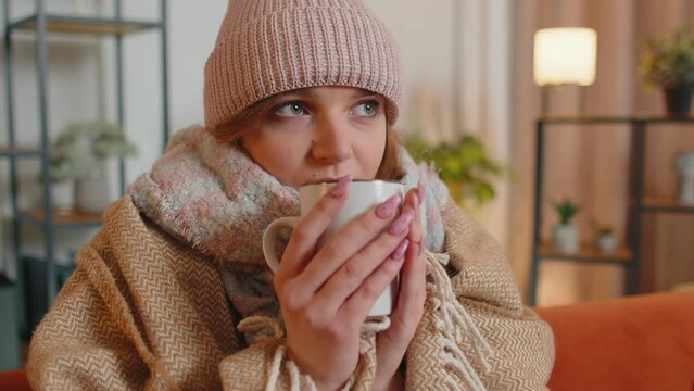 Young sick woman wear hat wrapped in plaid sit alone shivering from cold on sofa drinking hot tea in unheated apartment without heating due debt. Unhealthy female feeling discomfort try to warming up
