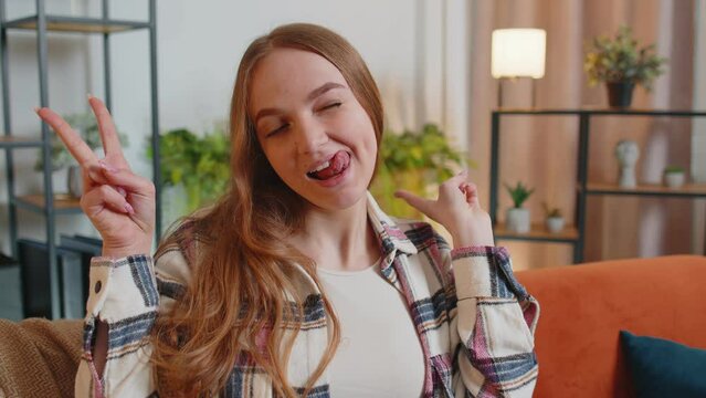 Funny amusing young funny woman in shirt looking awkward making playful silly facial expressions and grimacing, fooling around, showing tongue making stupid brainless dumb expression indoors at home