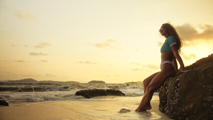 Silhouette sexy woman leaning on a rock reef enjoying and relax warm dark golden sunset. Woman in...