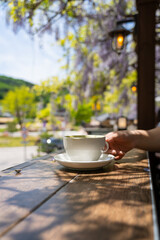 Woman hand holding a Hot coffee in a white ceramic cup tea set with purple wisteria flowers hanging at the outdoor cafe in South Korea