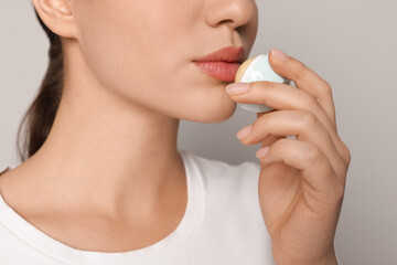 Young woman applying lip balm on grey background, closeup