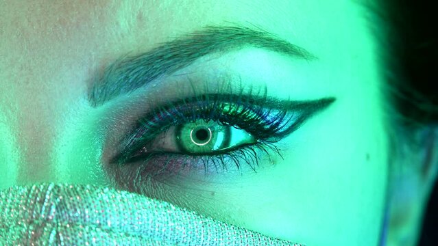 Woman eye with fancy arrow make-up under glowing neon light. Lady in protective mask. Glitter shadows and false lashes. Girl, green eye. Pandemic, covid-19 concept.