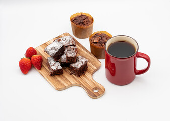 Brownies on wooden board, chocolate muffins, assorted chocolates on white plate, red coffee cup, strawberries and white background