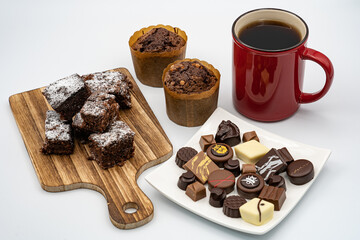 Brownies on wooden board, chocolate muffins, assorted chocolates on white plate, red coffee cup and white background