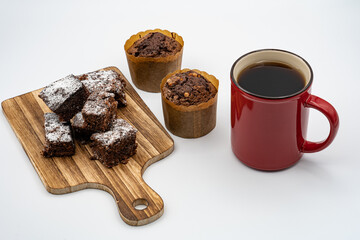 Brownies on wooden board, chocolate muffins, assorted chocolates on white plate, red coffee cup and white background