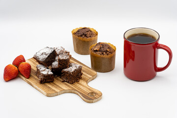 Brownies on wooden board, chocolate muffins, assorted chocolates on white plate, red coffee cup, strawberries and white background