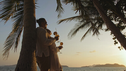 SILHOUETTE DARK GOLD SUNSET VIEW: Loving couple in white shirt and sunglasses, near palm tree, kissing, hugging, relax and drinking cocktail. Concept tropical summer vacations, holidays wedding love
