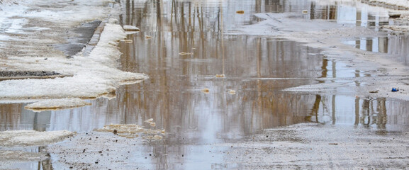 A road with melting snow, puddles and ice on a cloudy spring day. Reflections in cold water. Snow...