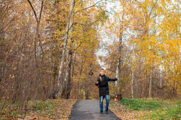 Obraz premium A teenage boy walks through an autumn park and shoots a video. A Caucasian boy in a black jacket holds a monopod with a smartphone. Blogger. Emotional communications..