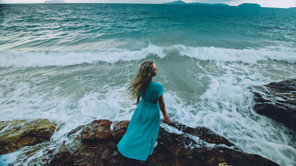 Sexy hot woman meditates, relaxes on a rock reef hill in stormy morning rain cloudy sea. Girl in blue swimsuit, dress tunic. Concept feminine, relax, sexual health. Dark dramatic silhouette view