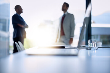 Big talk in the boardroom. Shot of an open laptop on a boardroom desk with two businessmen blurred in the background.