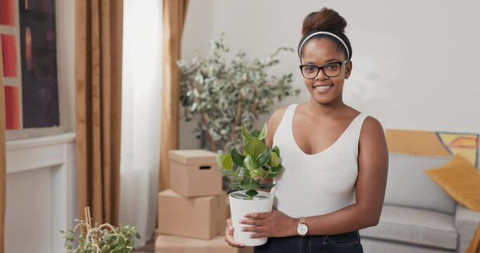 Smiling girl is standing in middle of living room in new rented purchased apartment, decorating house after moving, cleaning, in her hands holding a plant in a pot to be placed on the windowsill
