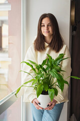 Beautiful young caucasian woman holding a home plant Spathiphyllum in a pot. Gardening, hobby. Soft selective focus.