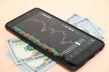 Stock exchange chart on smartphone screen and hundred dollar bills on beige background.